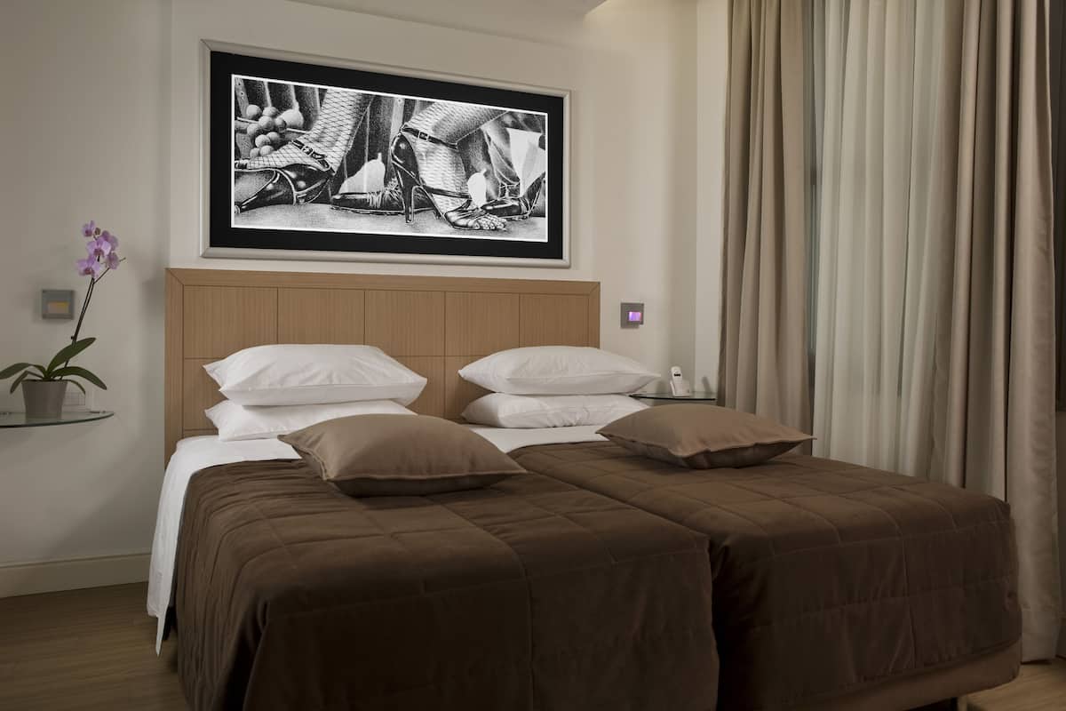 A hotel room with a double bed, two pillows, a framed artwork above the bed, a bedside table with a plant, and a window with curtains.