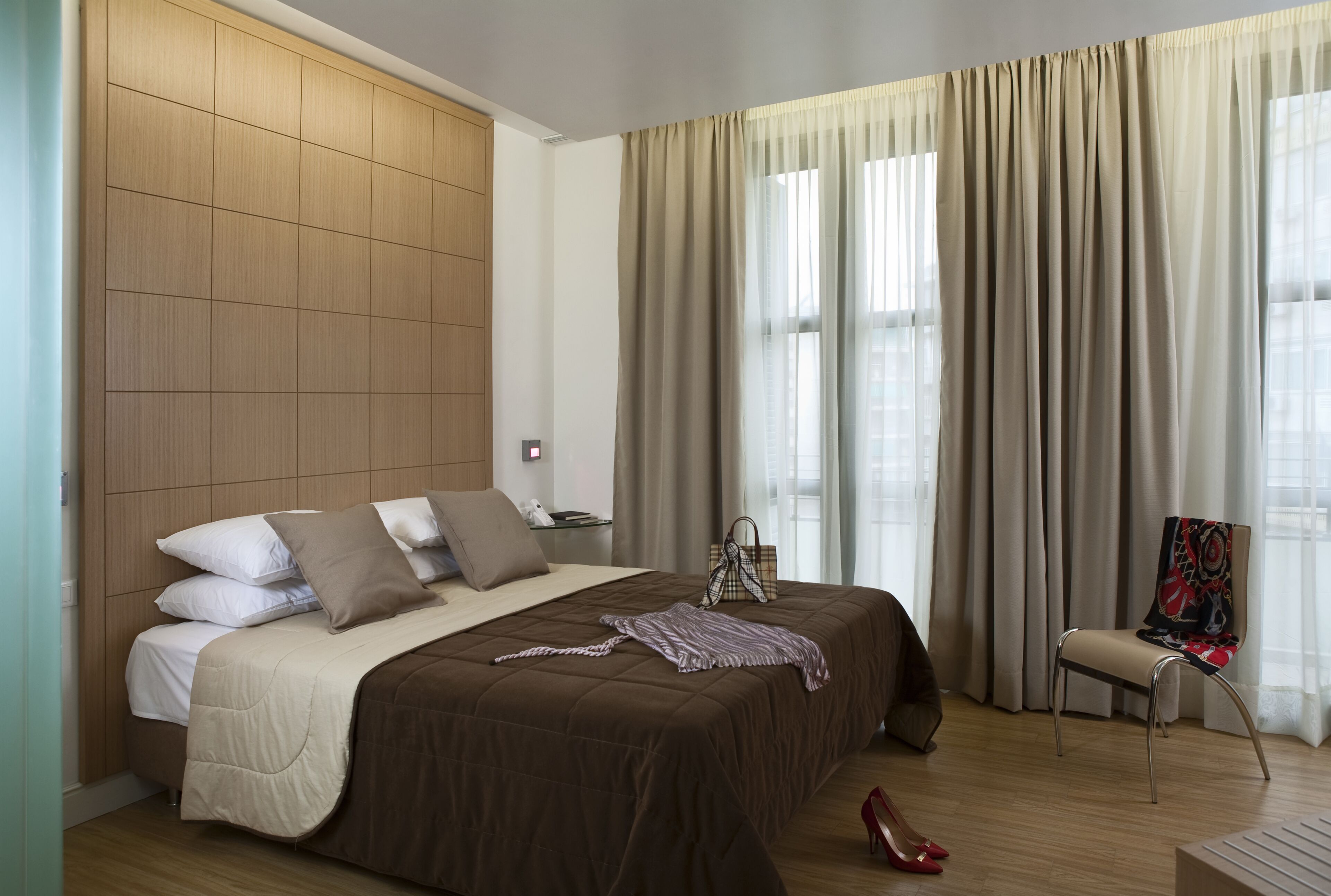 Habitación de hotel moderna con una cama grande, cabecero de madera, una silla y vistas a la ciudad a través de amplios ventanales.
