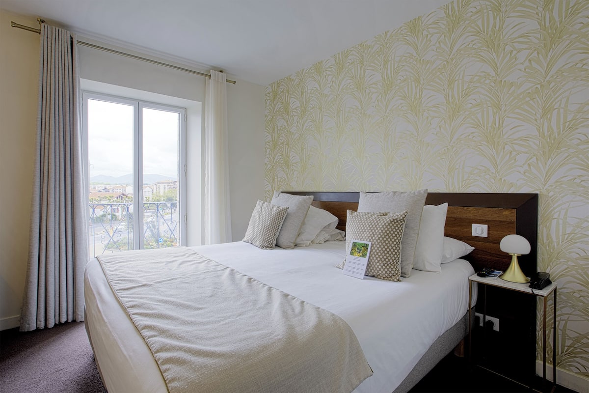 A hotel room with a large bed, a nightstand with a lamp, and a view of the city through a window.