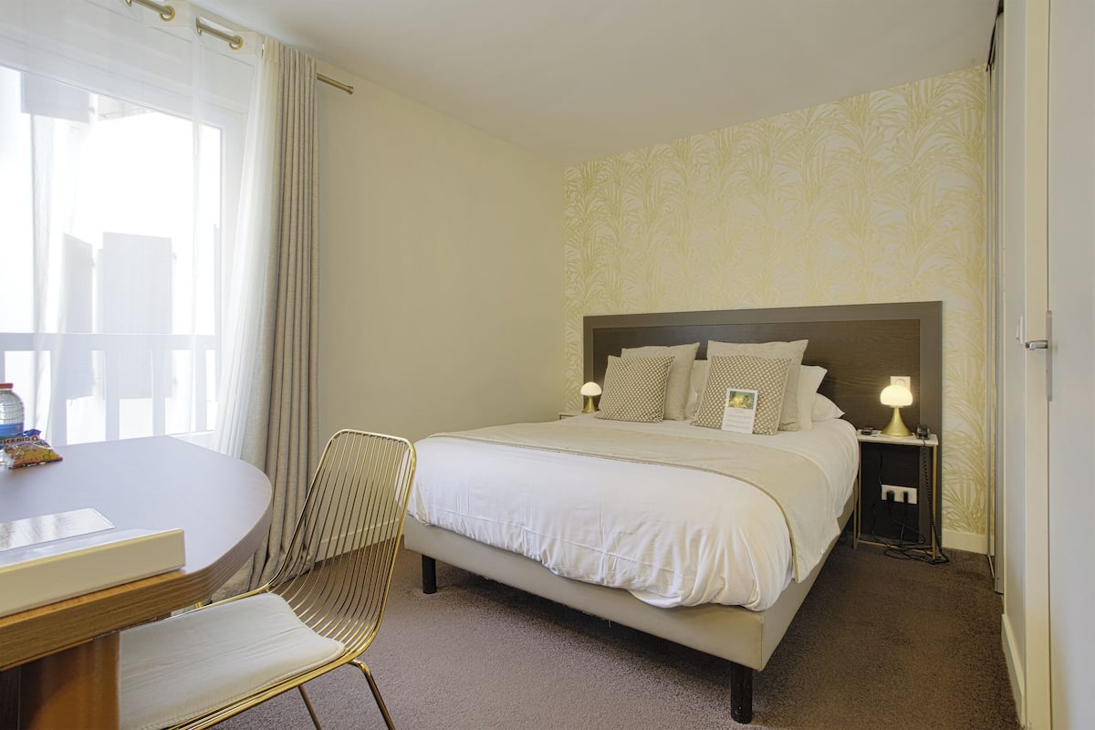 A hotel room with a bed, bedside table, lamp, chair, and a view of the outside through a window.