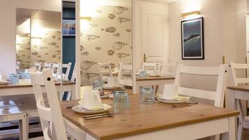 Un comedor con sillas blancas y una mesa de madera, decorado con papel tapiz estampado con motivos de peces y un cuadro en la pared.