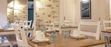 Un comedor con sillas blancas y una mesa de madera, decorado con papel tapiz estampado con motivos de peces y un cuadro en la pared.