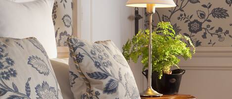 Una cama bien hecha con almohadas de estampado floral, una lámpara de noche y una planta en maceta sobre una mesita de madera.