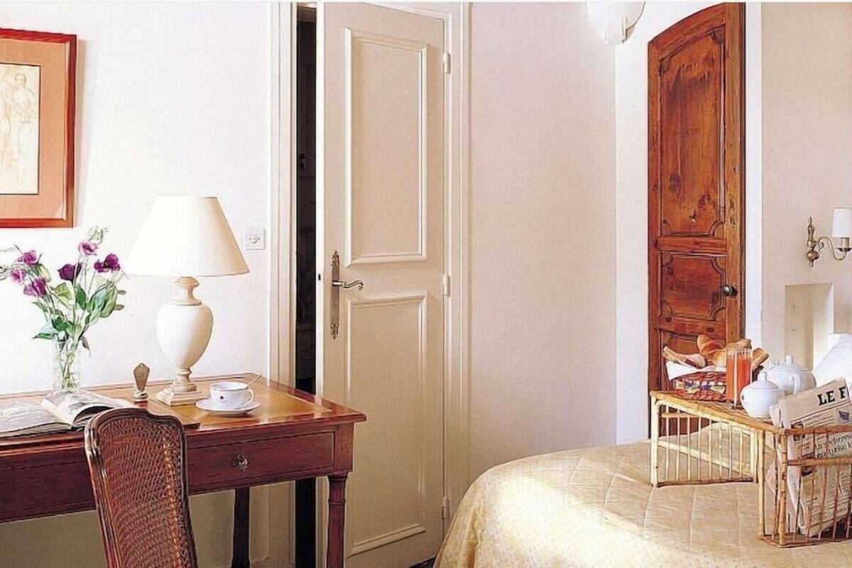 A hotel room with a wooden desk, a chair, a bed with a headboard, a bedside table with a lamp, and a vase with flowers.