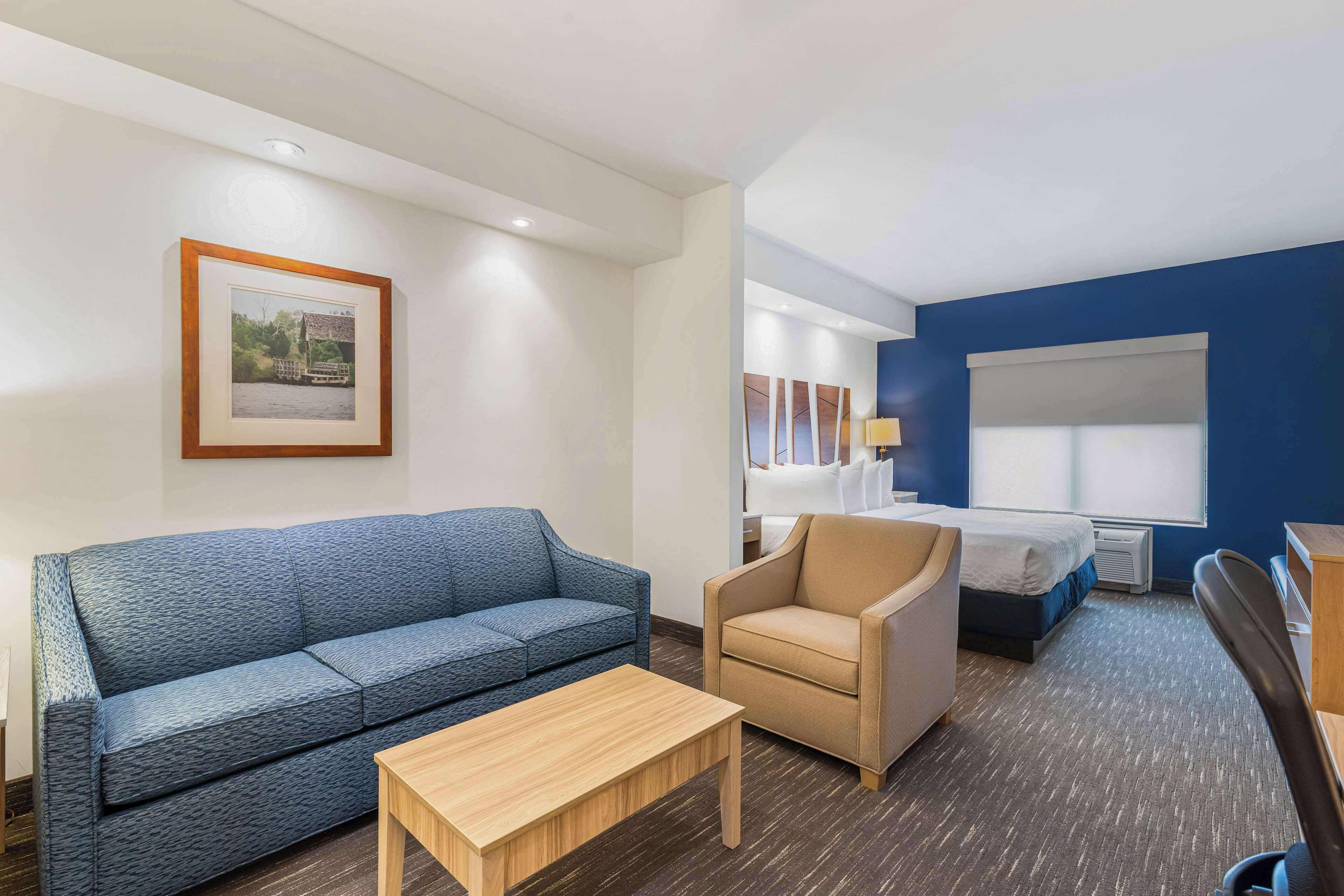 Ein Hotelzimmer mit einem blauen Sofa, einem beigen Sessel, einem Holztisch, einem Bett mit blauer Kopfteil und einem gerahmten Bild an der Wand.