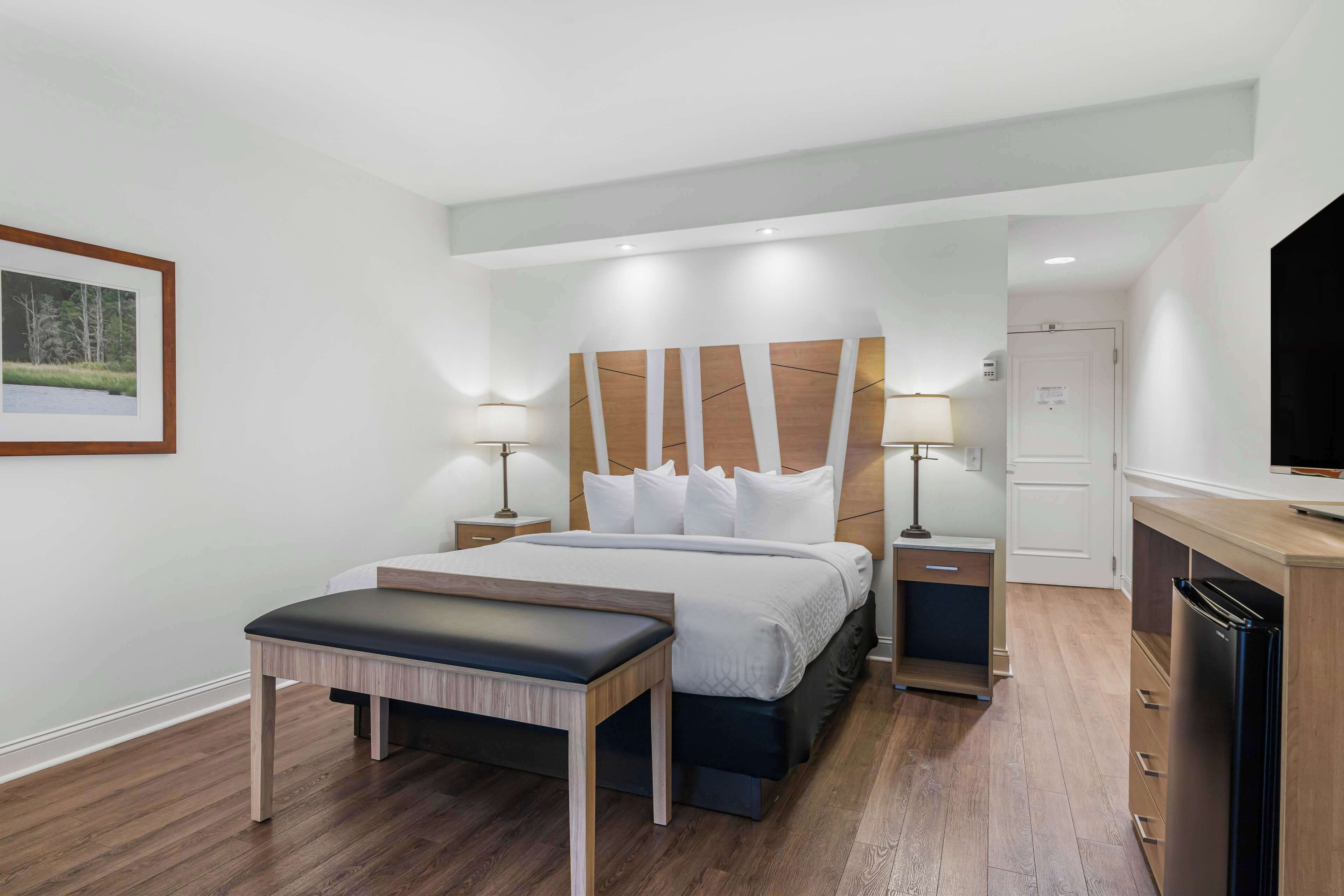 Una habitación de hotel moderna con una cama grande, mesitas de noche, un banco, un televisor y un refrigerador.