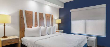 A hotel room with a large bed, two bedside tables with lamps, a window with blinds, and a wall-mounted air conditioner.