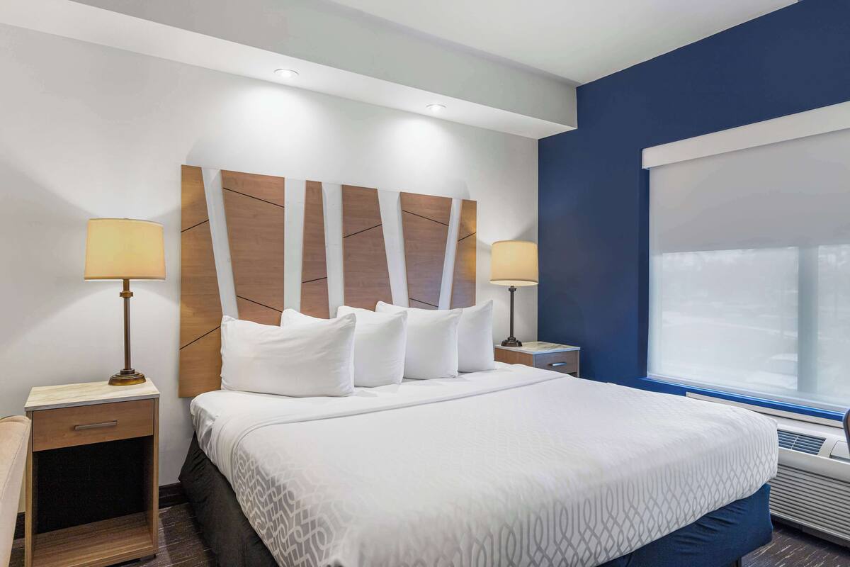 A hotel room with a large bed, two bedside tables with lamps, a headboard with a geometric design, a window with blinds, and an air conditioning unit.