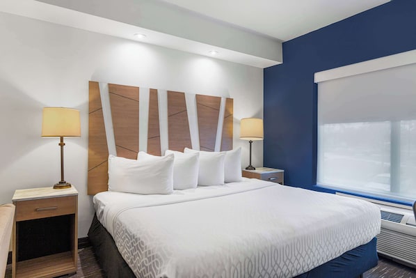 A hotel room with a large bed, two bedside tables with lamps, a headboard with a geometric design, a window with blinds, and an air conditioning unit.