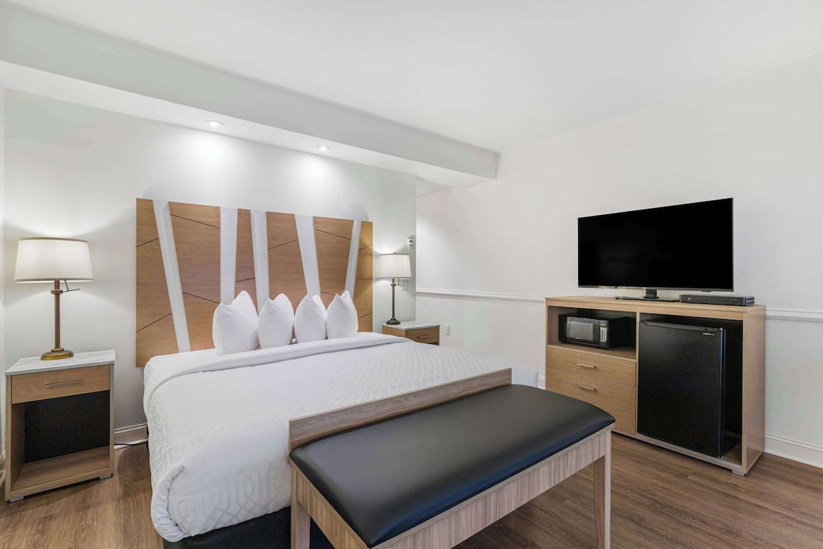 A modern hotel room with a large bed, bedside lamps, a flat-screen TV, a mini-fridge, and a wooden bench.