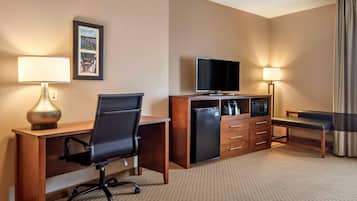 A hotel room with a wooden desk, a black office chair, a television, a mini-fridge, a coffee maker, and a bench.