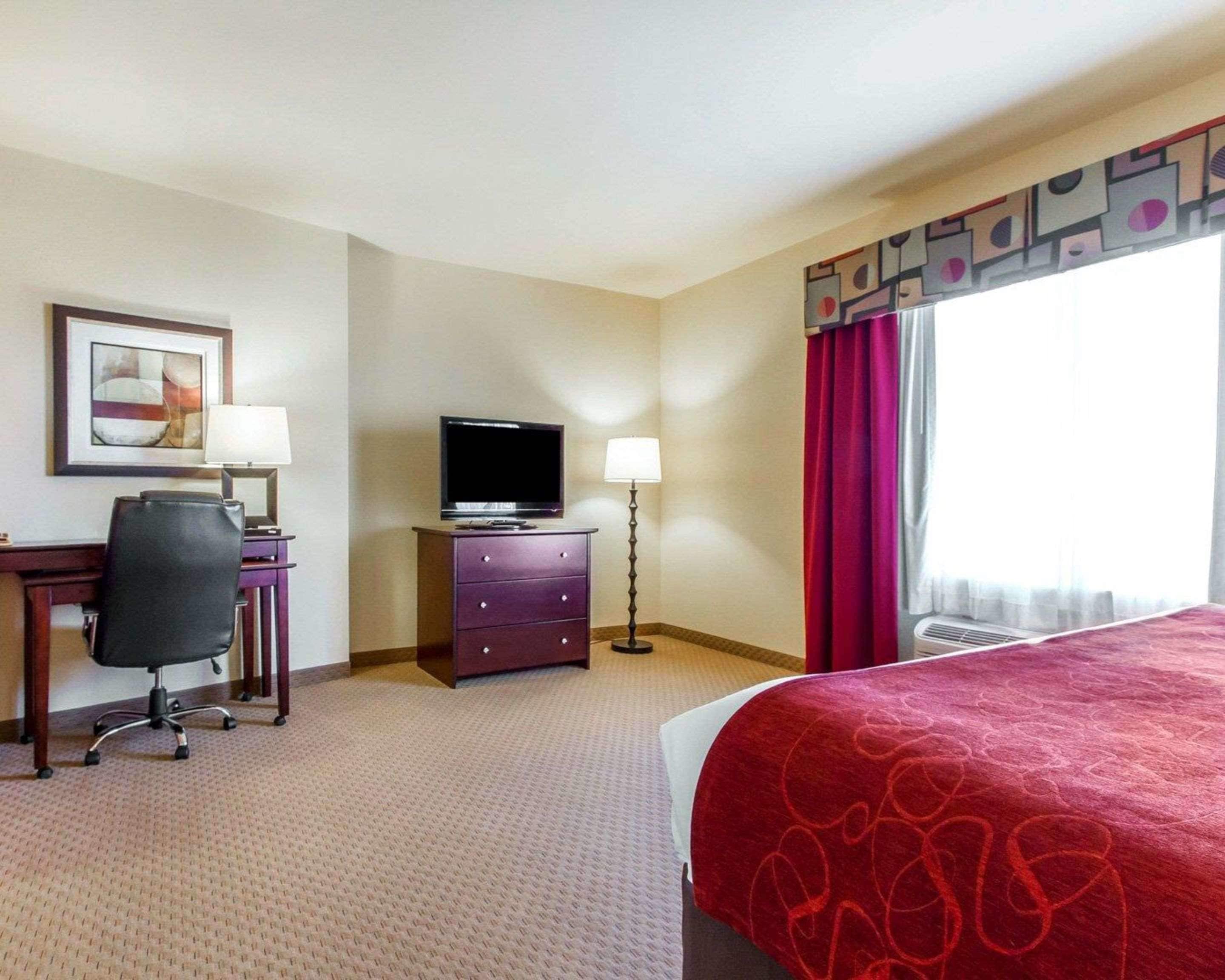 Habitación de hotel con cama, escritorio con silla, televisión, lámpara y ventana con cortinas.