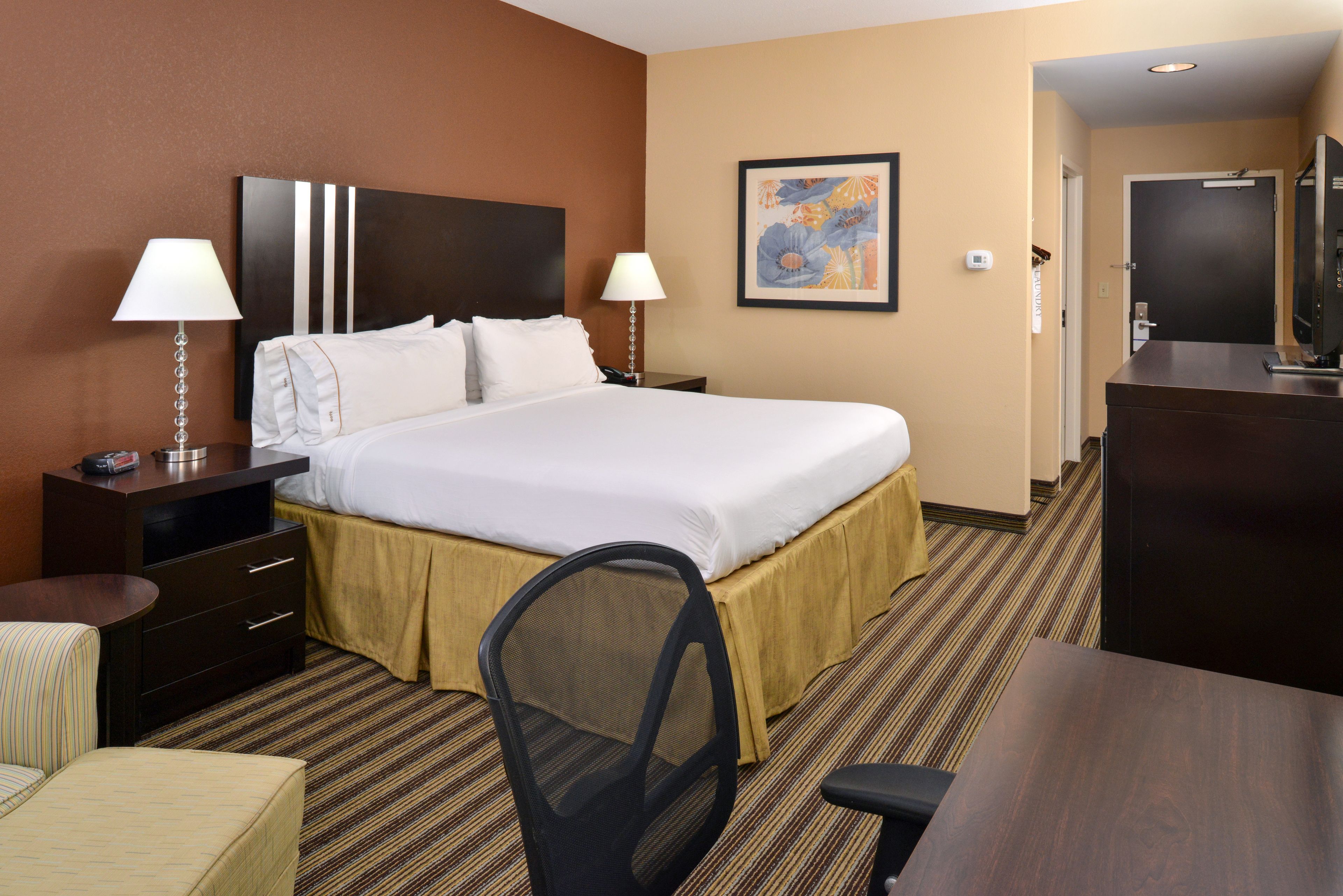 A hotel room with a large bed, two bedside tables with lamps, a desk with a chair, a dresser, and a painting on the wall.