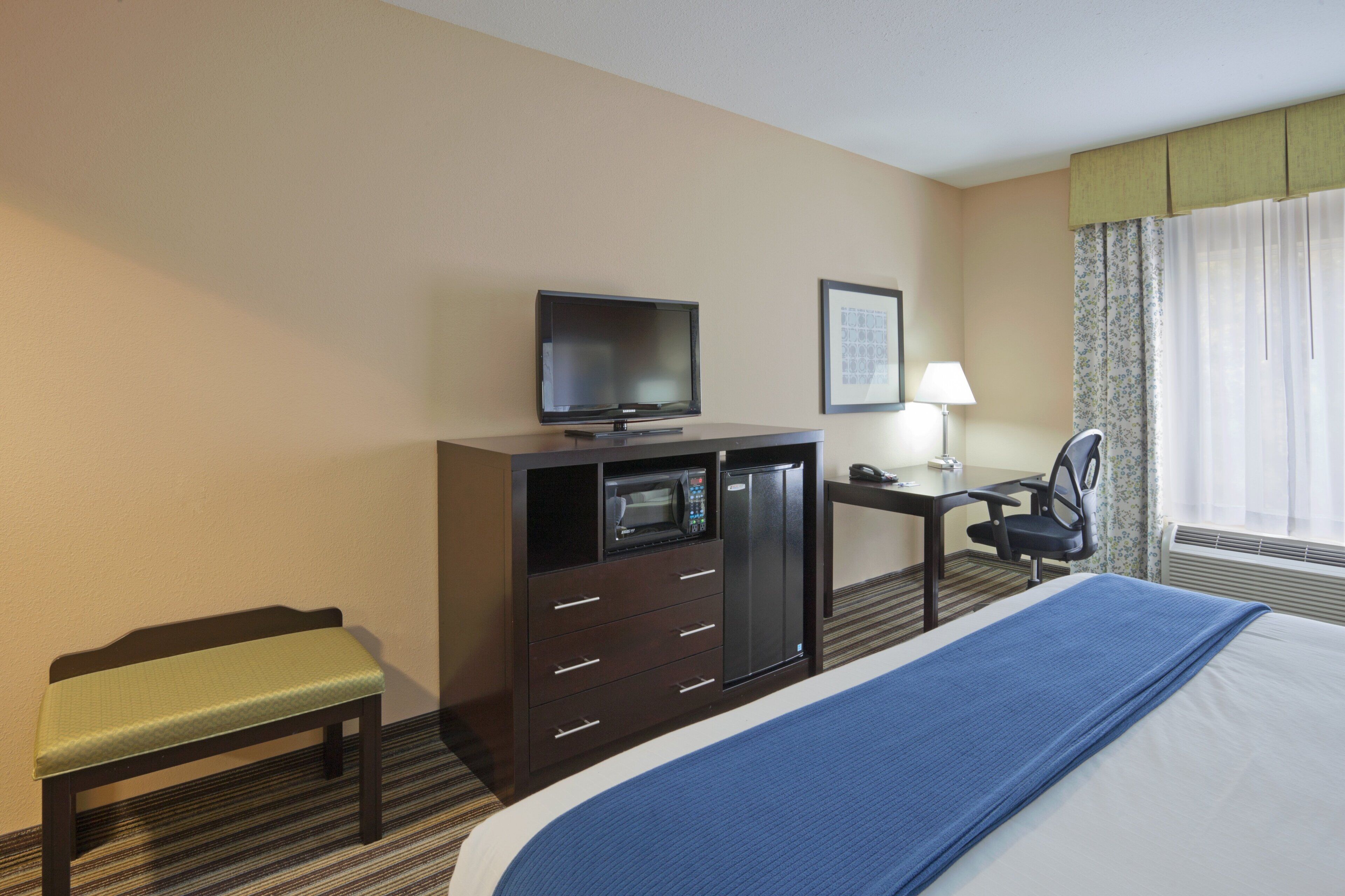 A hotel room with a bed, a desk with a lamp, a television, a microwave, a chair, and a bench.