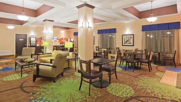 A hotel lobby with multiple seating areas, including sofas and chairs, arranged around a central column. There are tables and a counter with a menu display. The lobby features a patterned carpet and ceiling with recessed lighting. - Holiday Inn Express Columbia by IHG (Columbia)