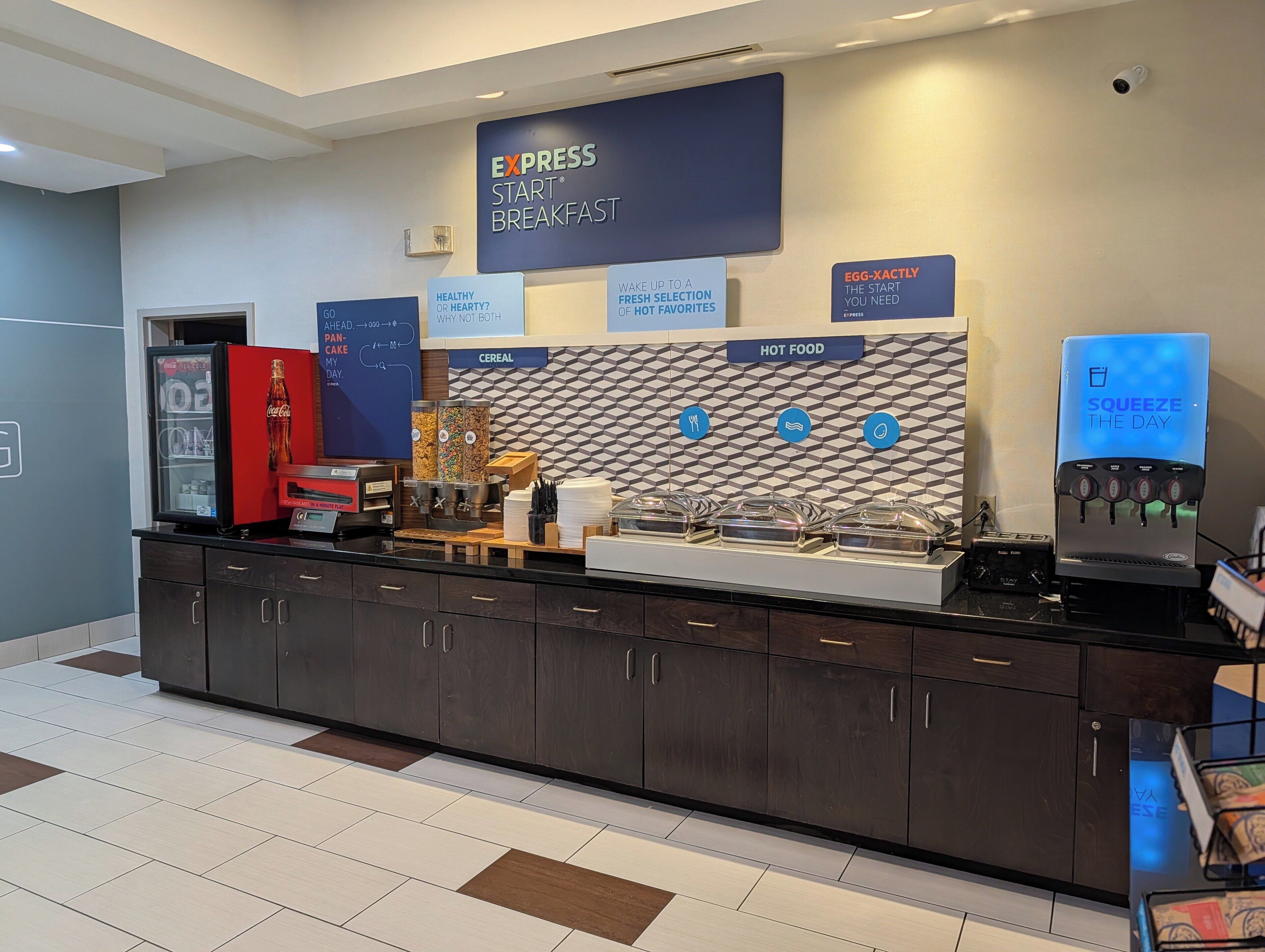 Zona de desayuno de hotel con autoservicio, un refrigerador de Coca-Cola y un letrero que anuncia el desayuno Express Start.
