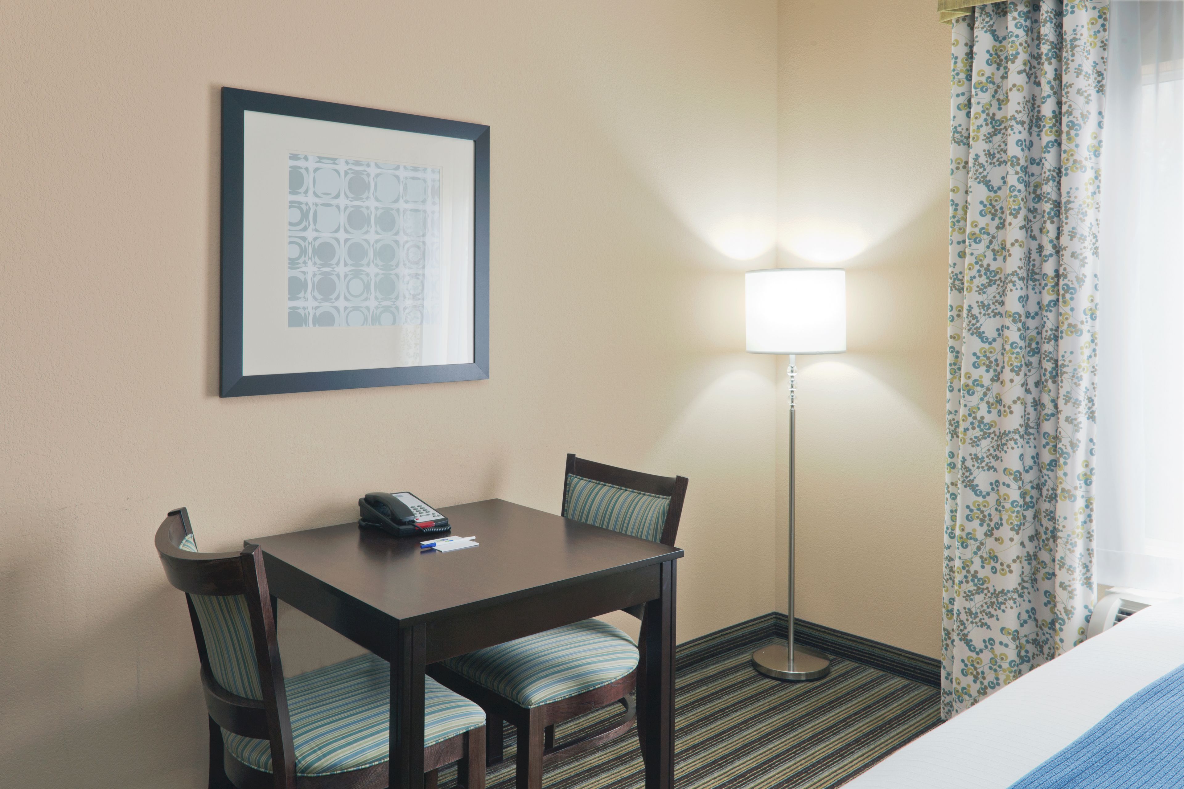 A hotel room with a bed, a desk with a telephone, a chair, a lamp, and a framed picture on the wall.