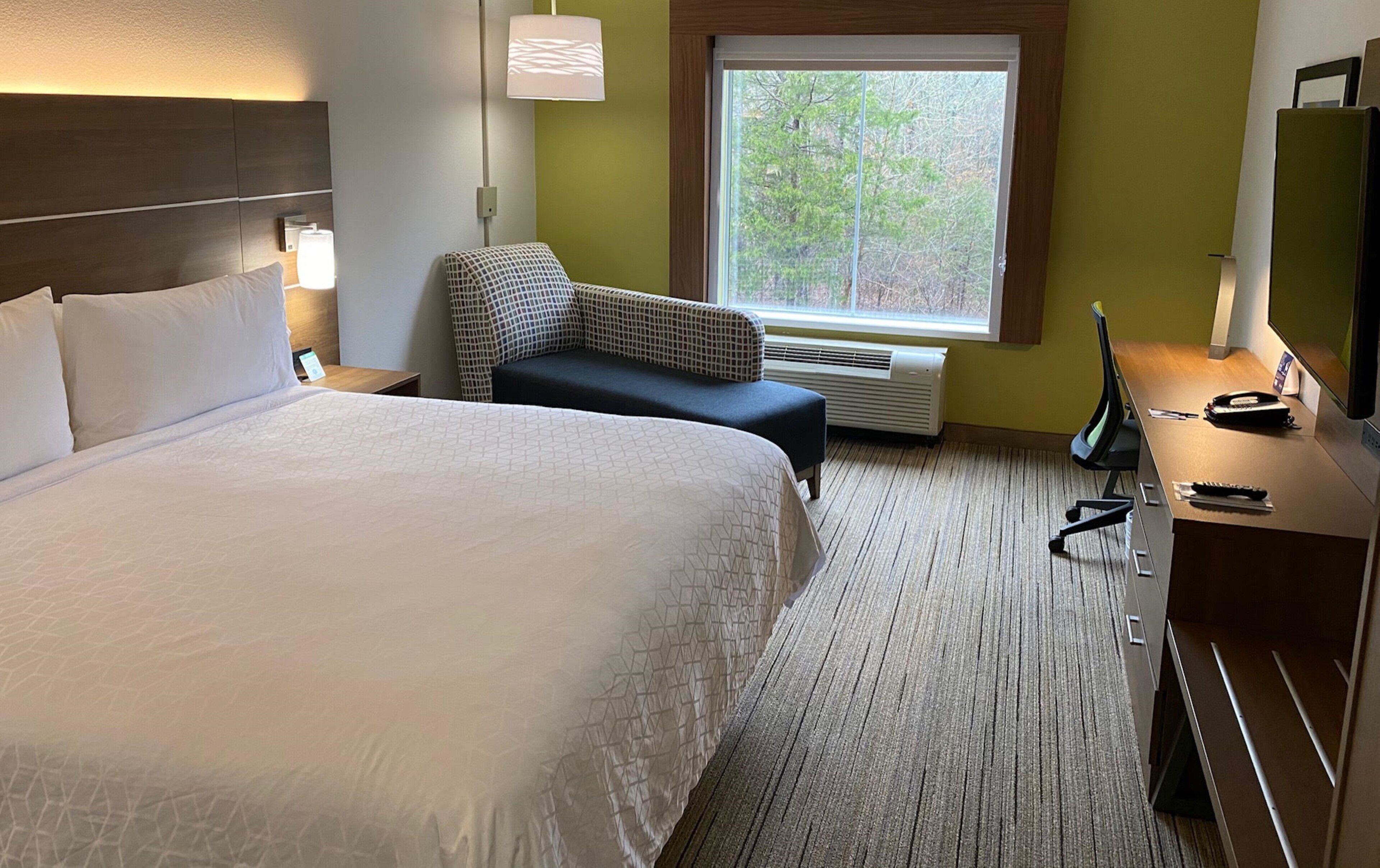 A hotel room with a large bed, a desk with a computer, a chair, and a window with a view of trees.
