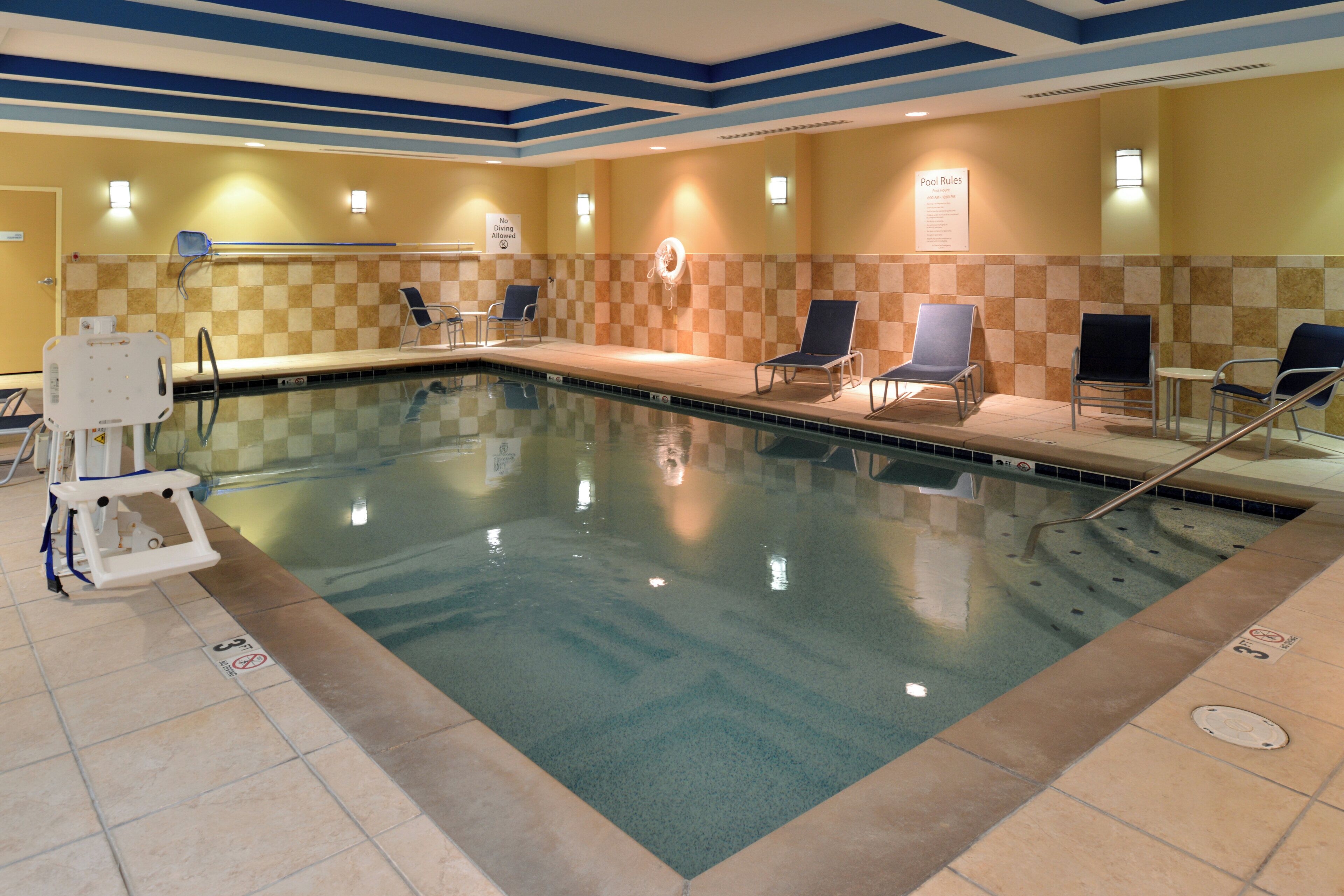 A swimming pool with lounge chairs and a lifeguard chair, surrounded by tiled walls and a blue ceiling.