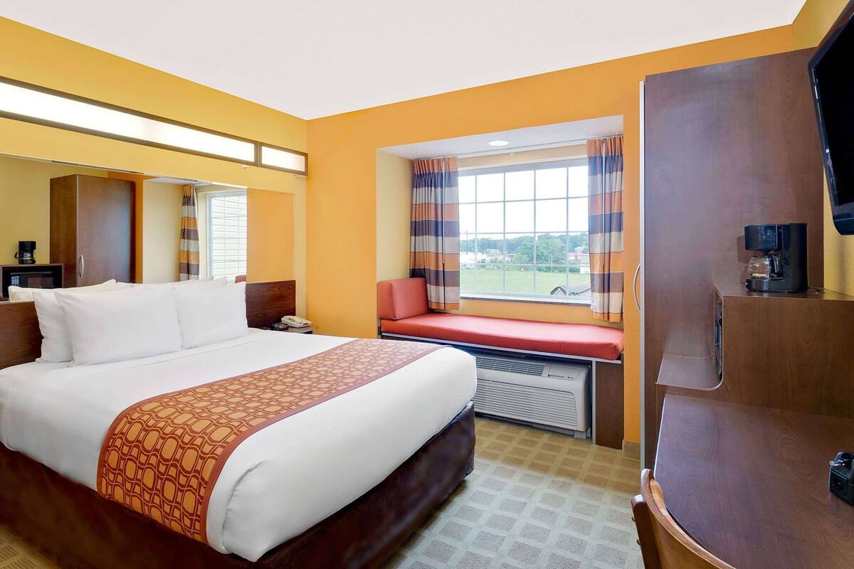 A hotel room with a bed, a desk with a computer, a window with curtains, a television, and a coffee machine.