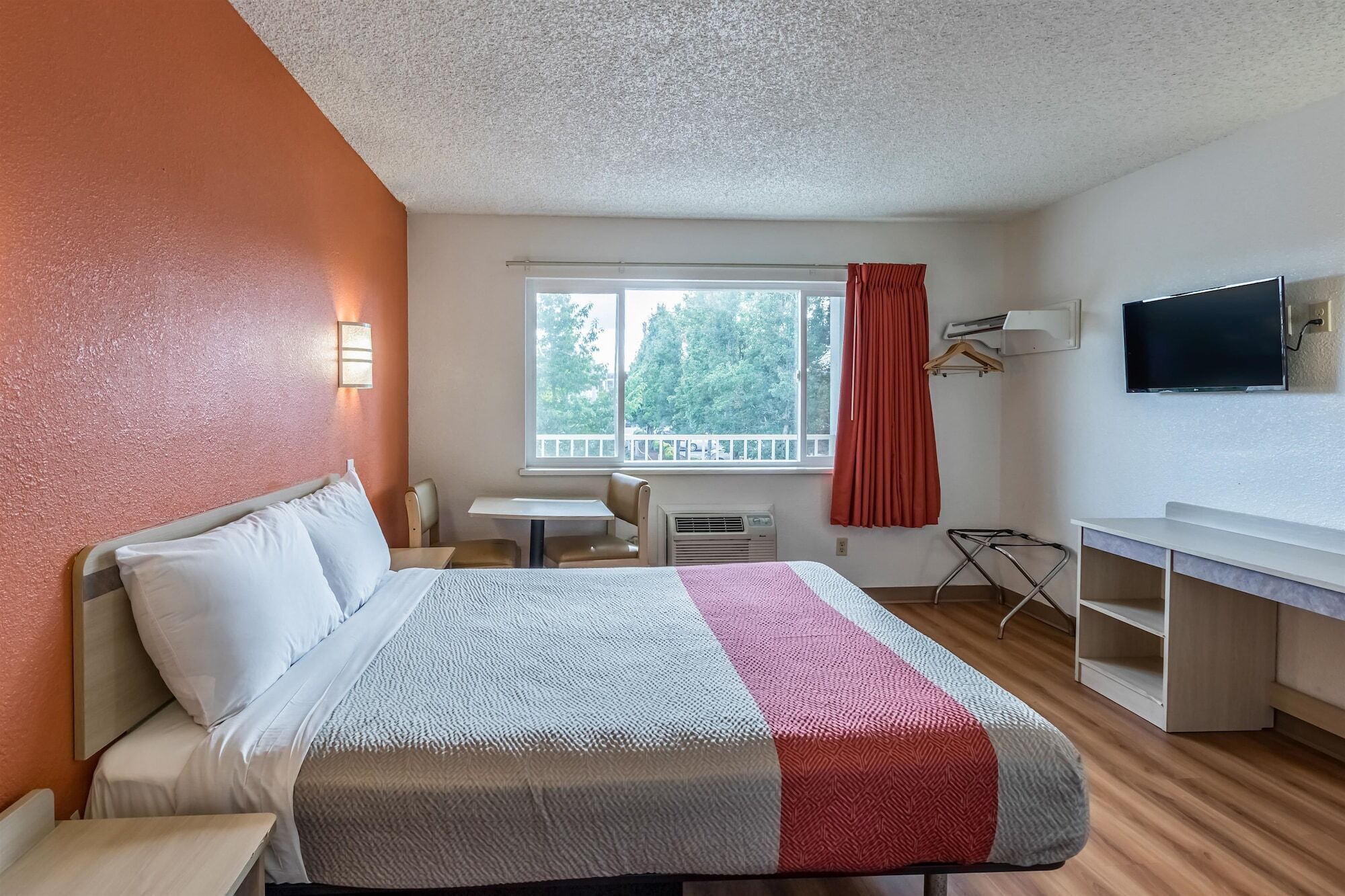 A hotel room with a bed, a desk, a chair, a TV, and a window with a view of trees.