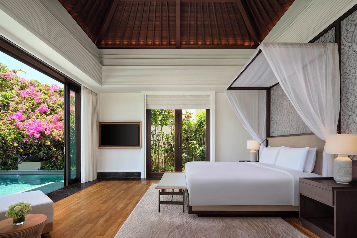 A bedroom with a bed, bedside tables, a TV, and a view of a swimming pool and garden.