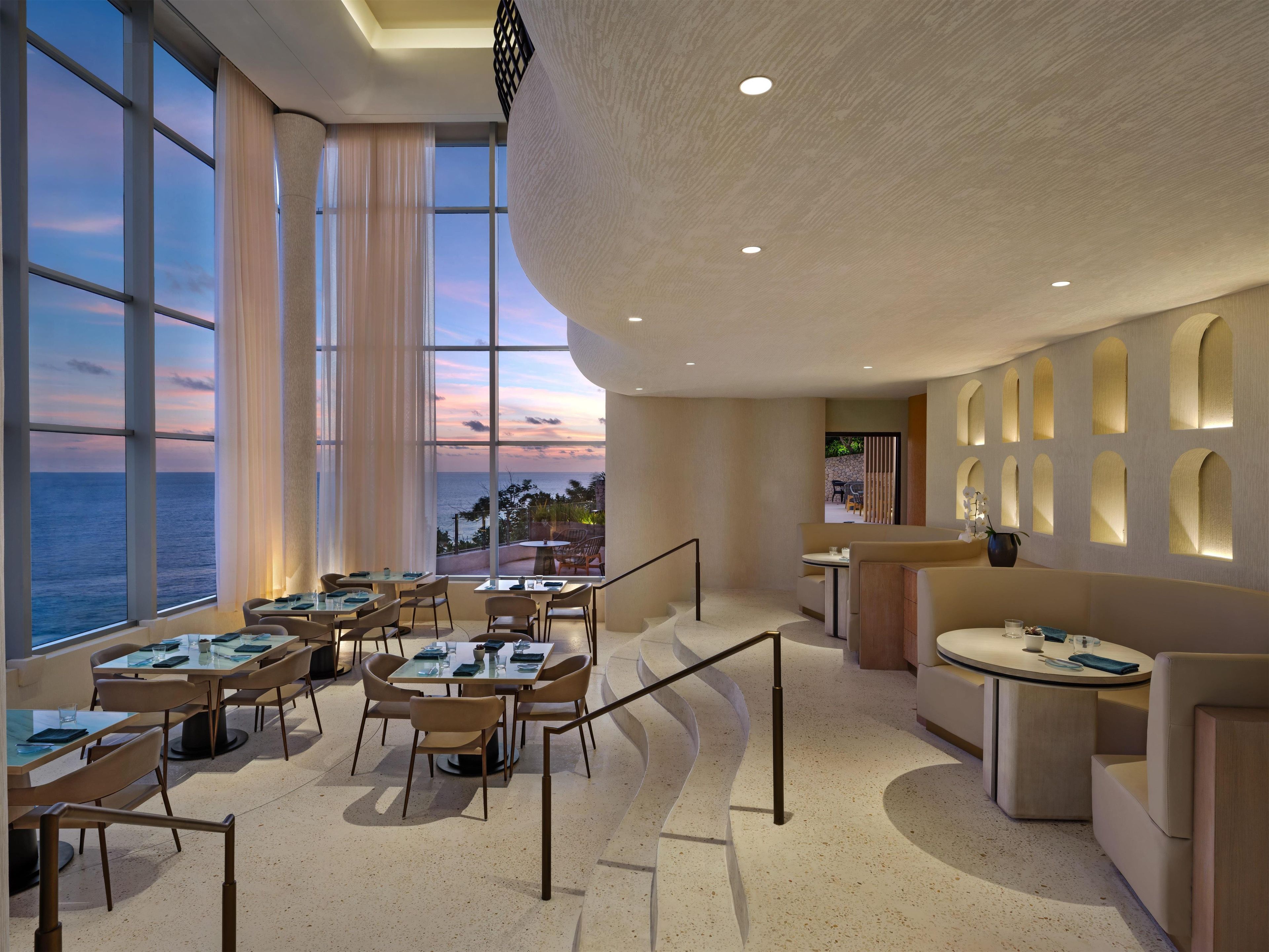 A modern dining area with tables and chairs, large windows with a view of the sea, and a curved ceiling.