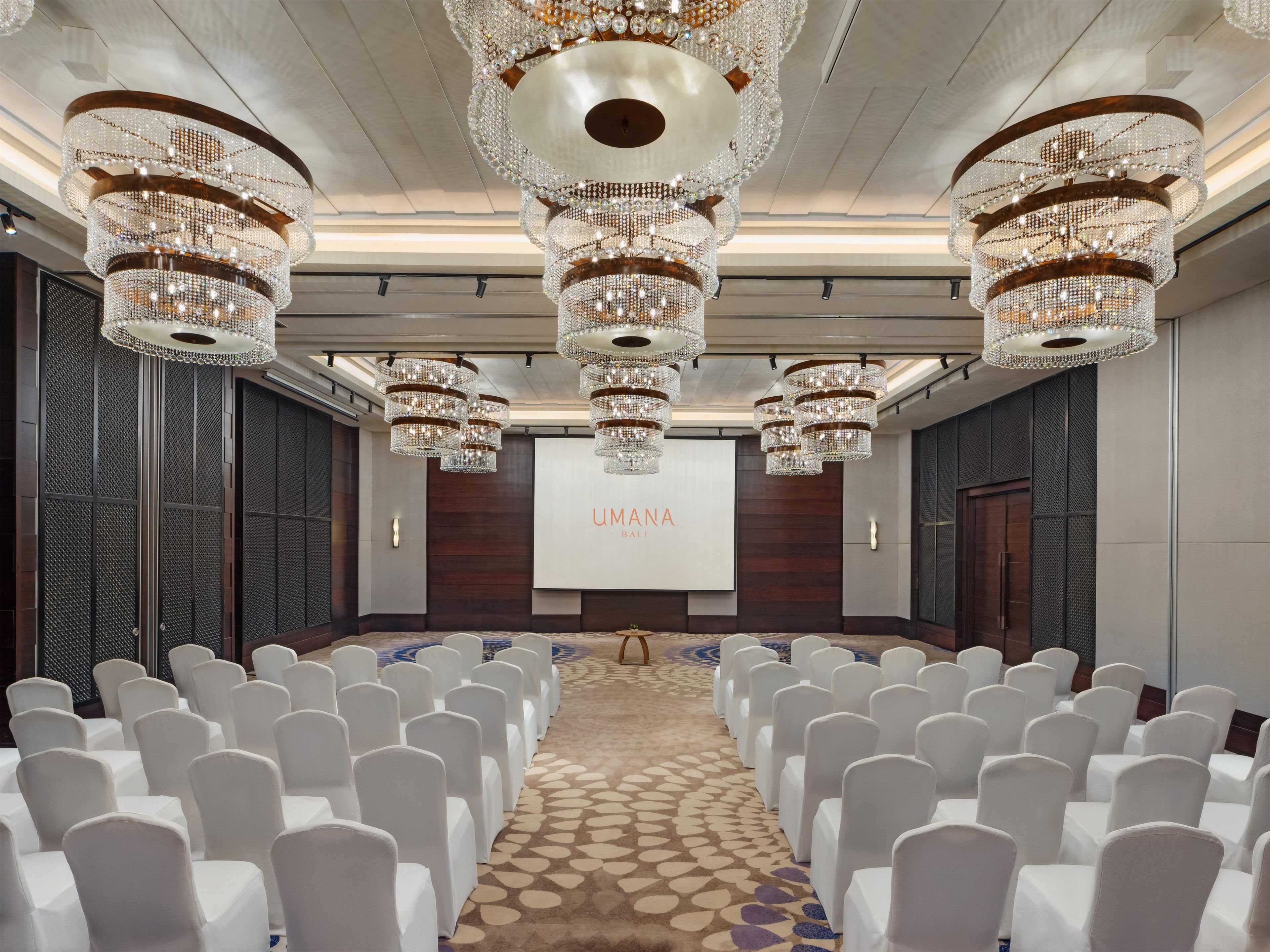 A conference room with rows of white chairs, a large screen displaying 'UMANA BALLI', and multiple chandeliers.