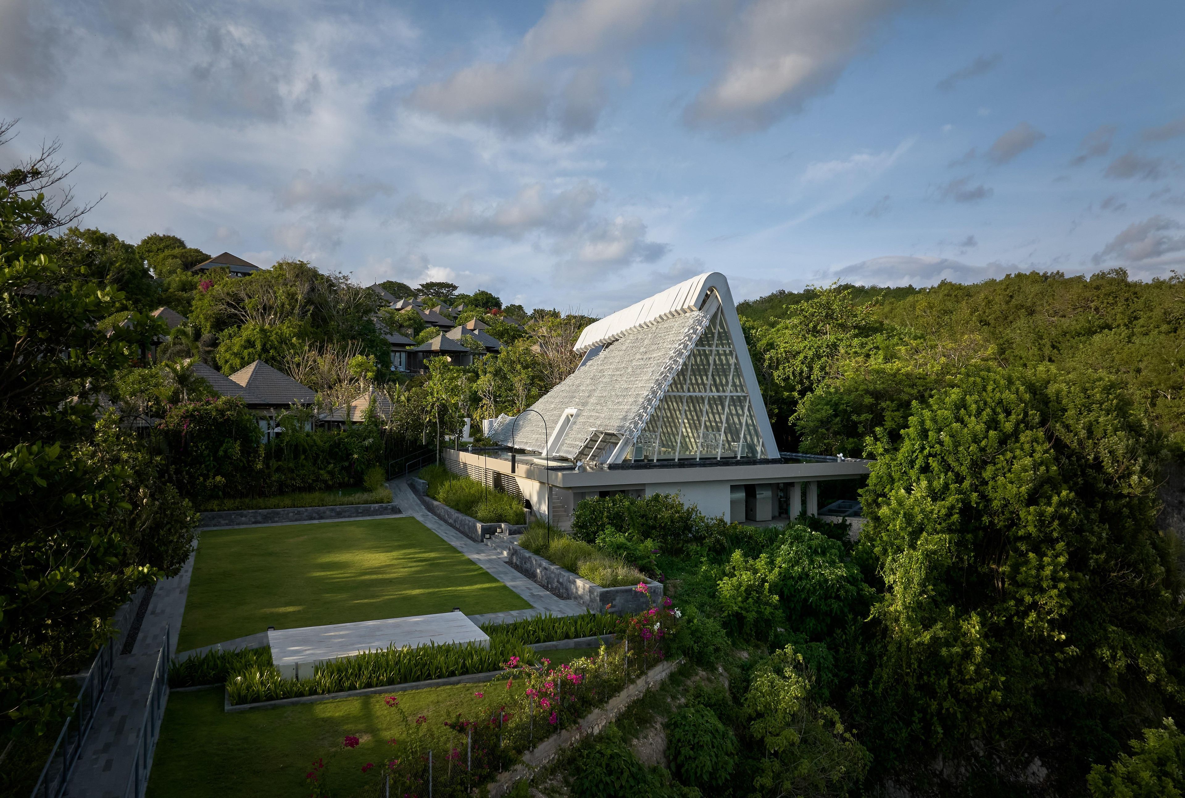 A modern building with a unique triangular roof, surrounded by lush greenery and residential houses.