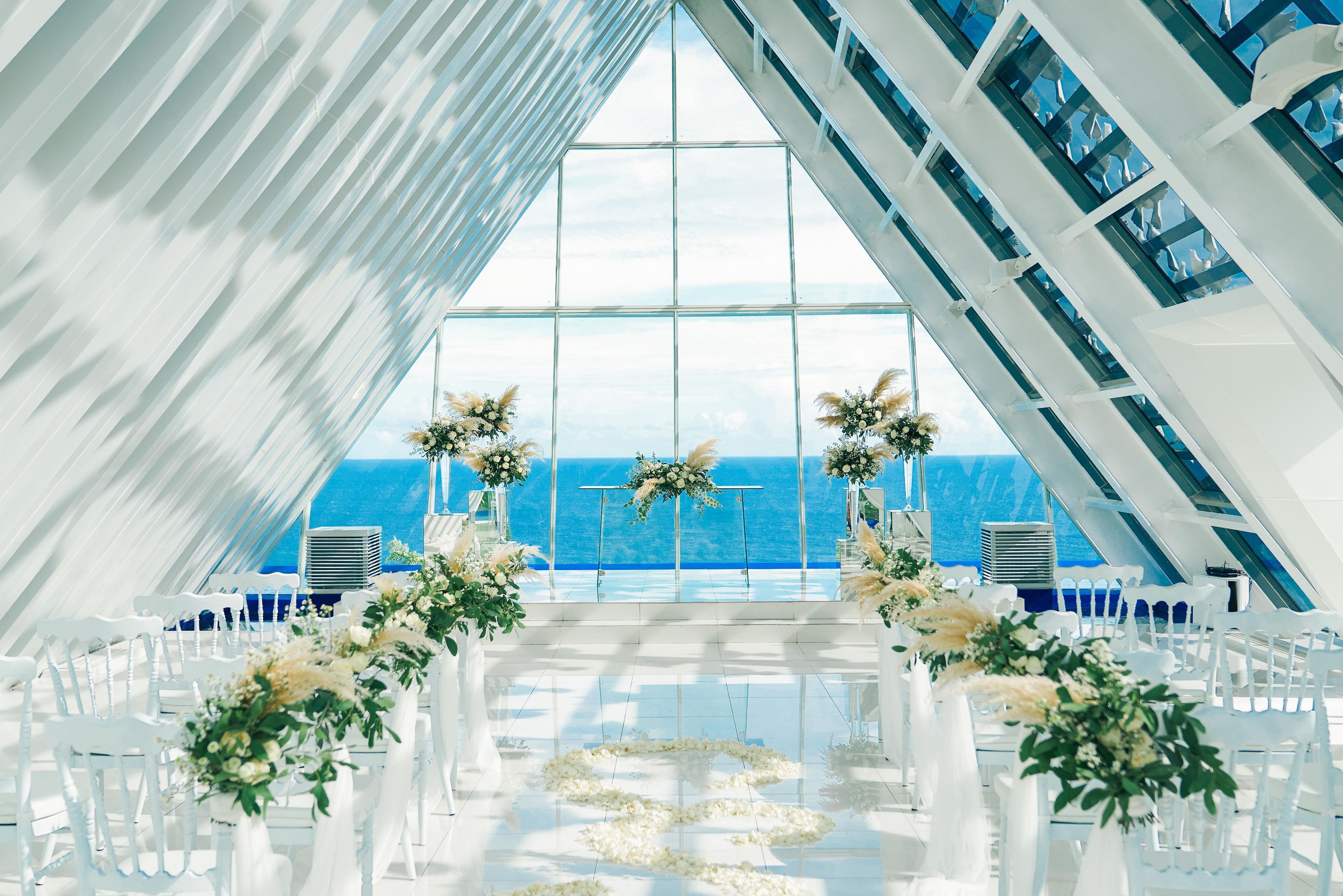 An indoor venue with a clear view of the ocean, white chairs arranged in rows, and floral decorations.
