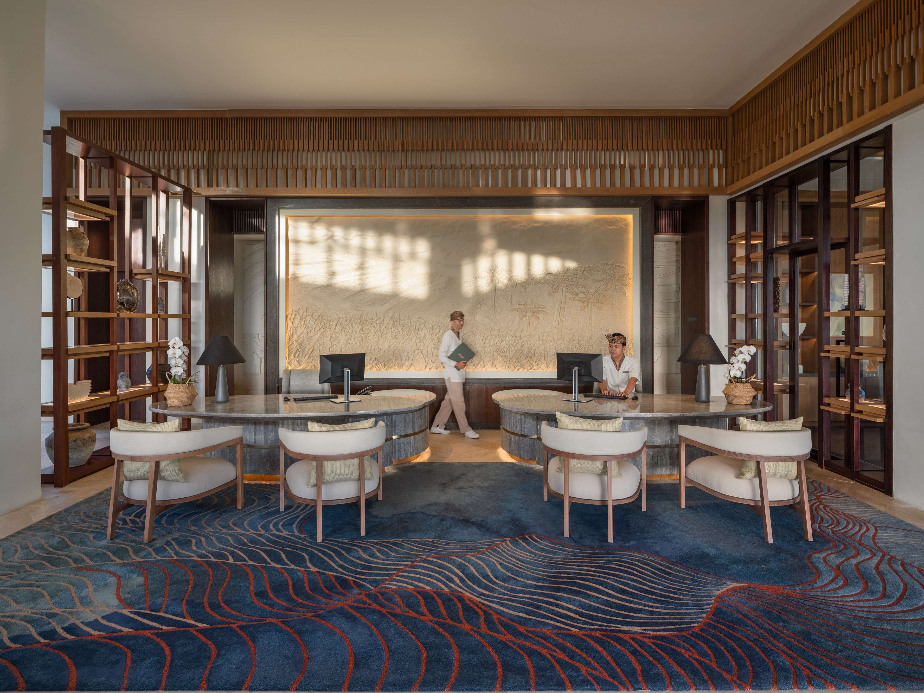 A modern hotel lobby with a central reception desk, two seating areas, and a large decorative wall.