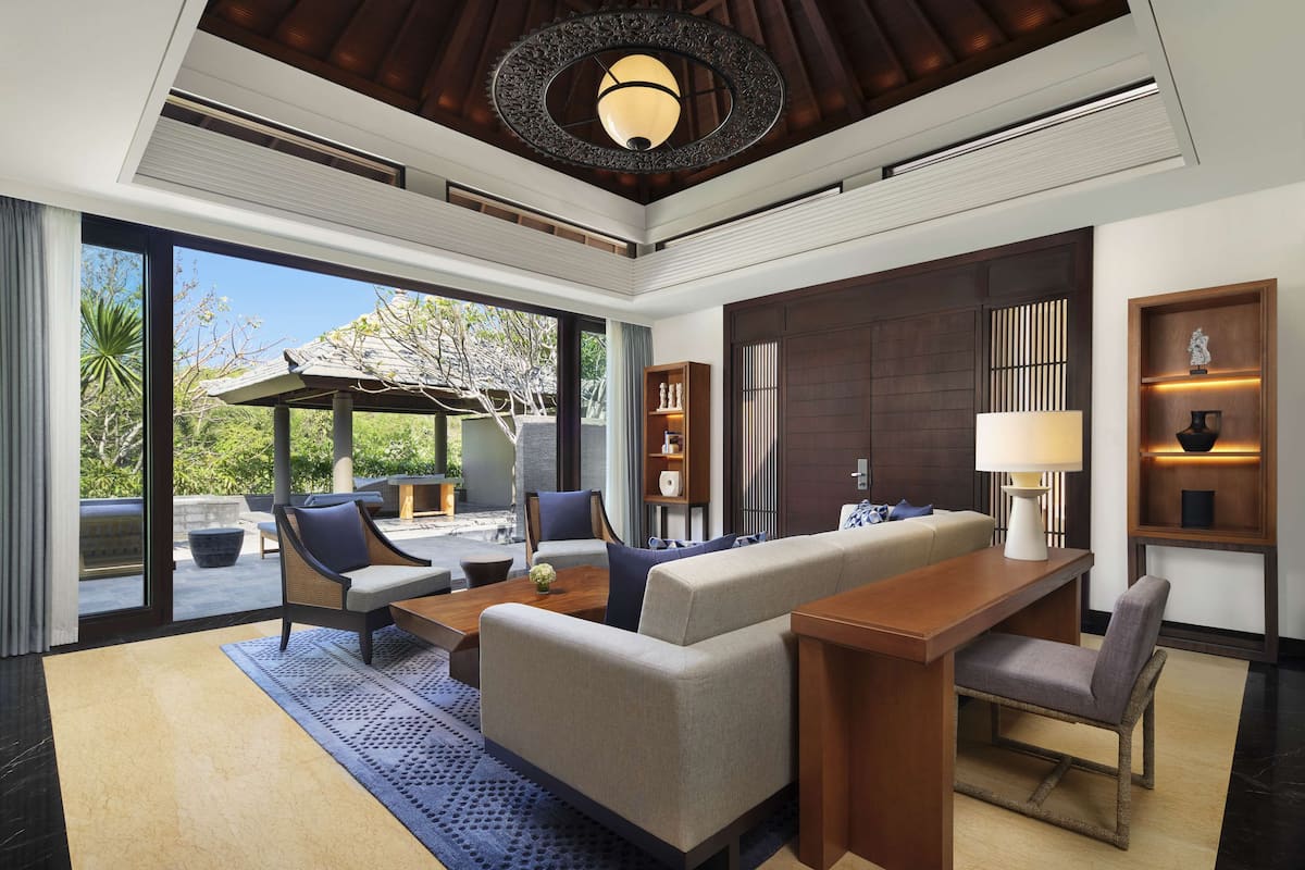 A modern living room with a sofa, chairs, a coffee table, and a dining table. There is a large window providing a view of the outdoors.