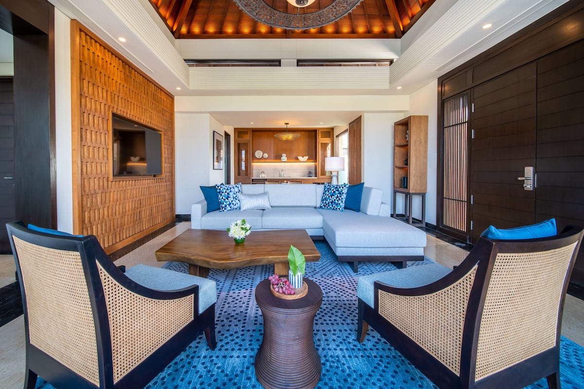 A modern living room with a sofa, coffee table, and wicker chairs. A kitchen is visible in the background.