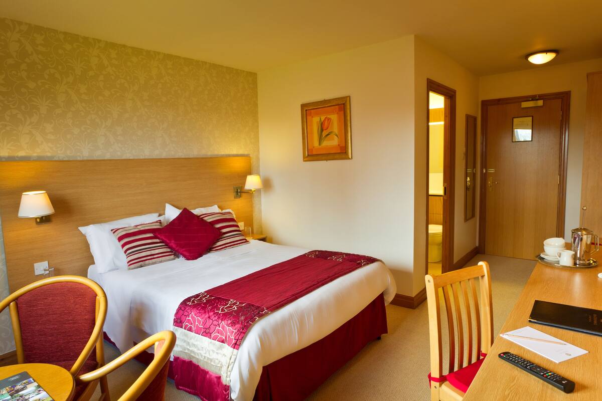Ein Hotelzimmer mit einem Bett, einem Schreibtisch mit Stuhl, einer Lampe, einem Bild an der Wand und einer Tür.