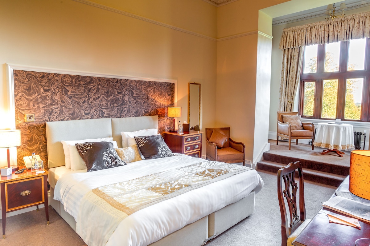 A hotel room with a large bed, a nightstand with a lamp, a chair, a table, and a window with curtains.