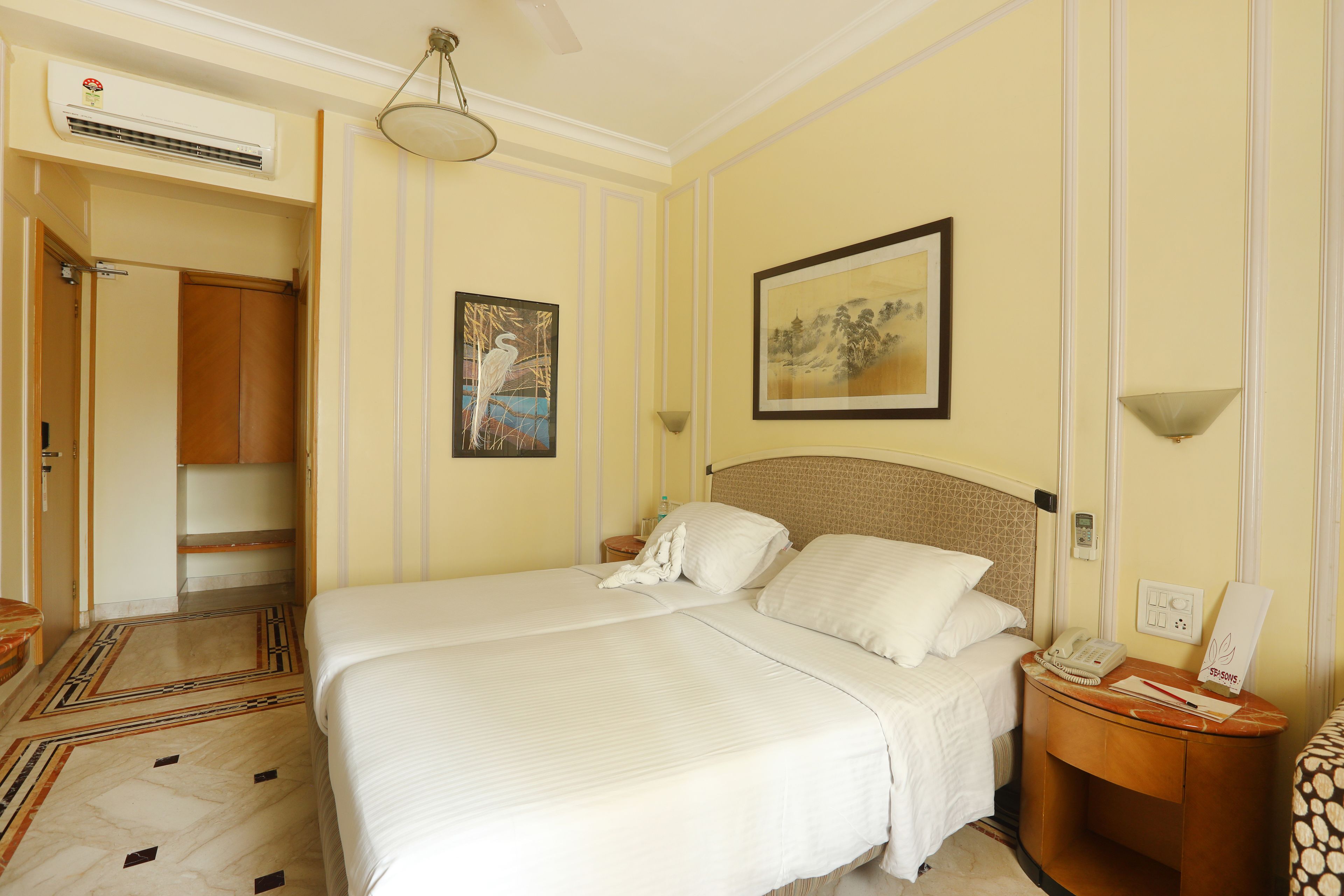 Una habitación de hotel con una cama grande, una mesita de noche con lámpara, un cuadro enmarcado en la pared y un equipo de aire acondicionado.