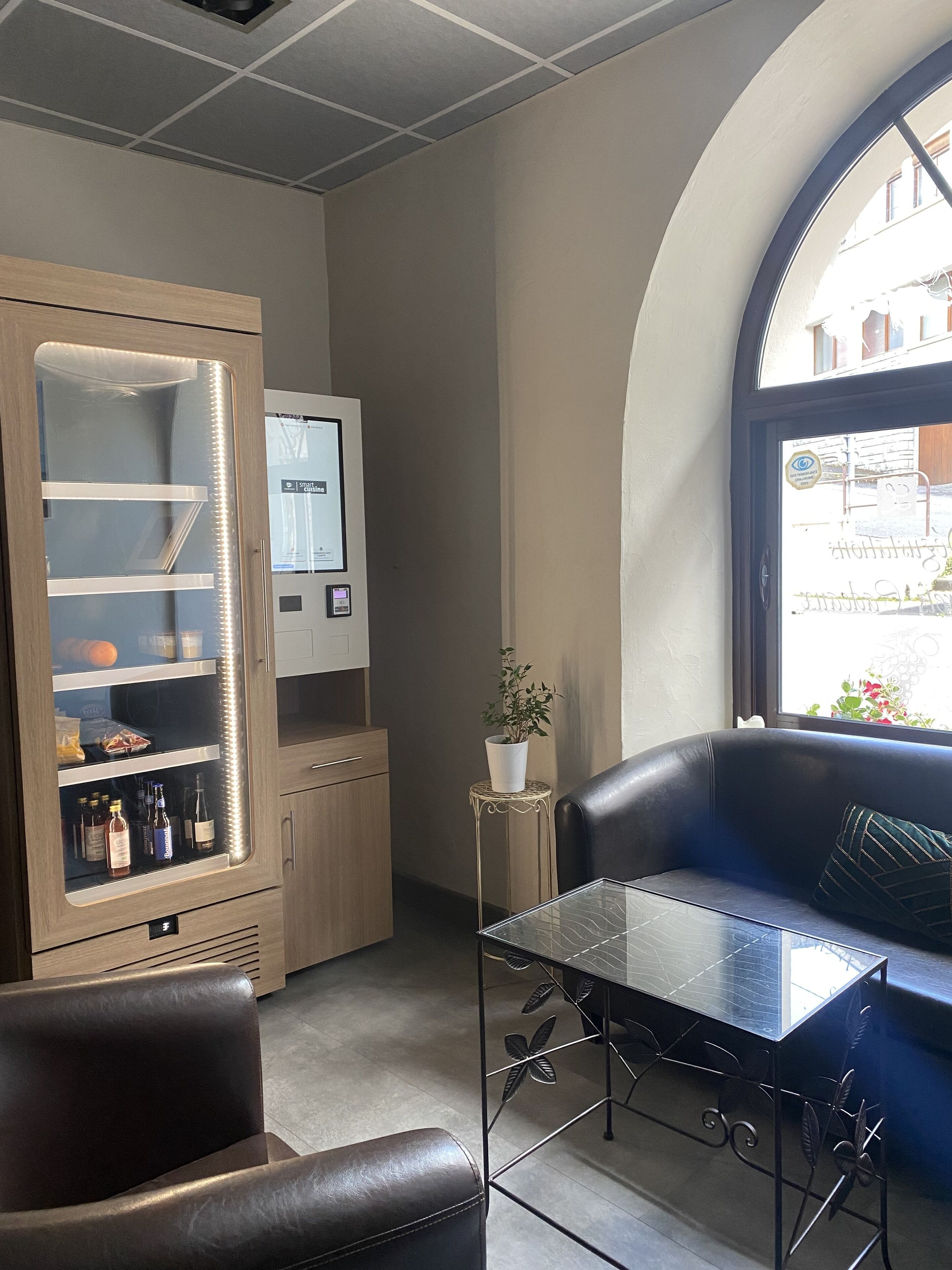 A modern interior with a seating area, a refrigerator with a digital display, and a view through a window.