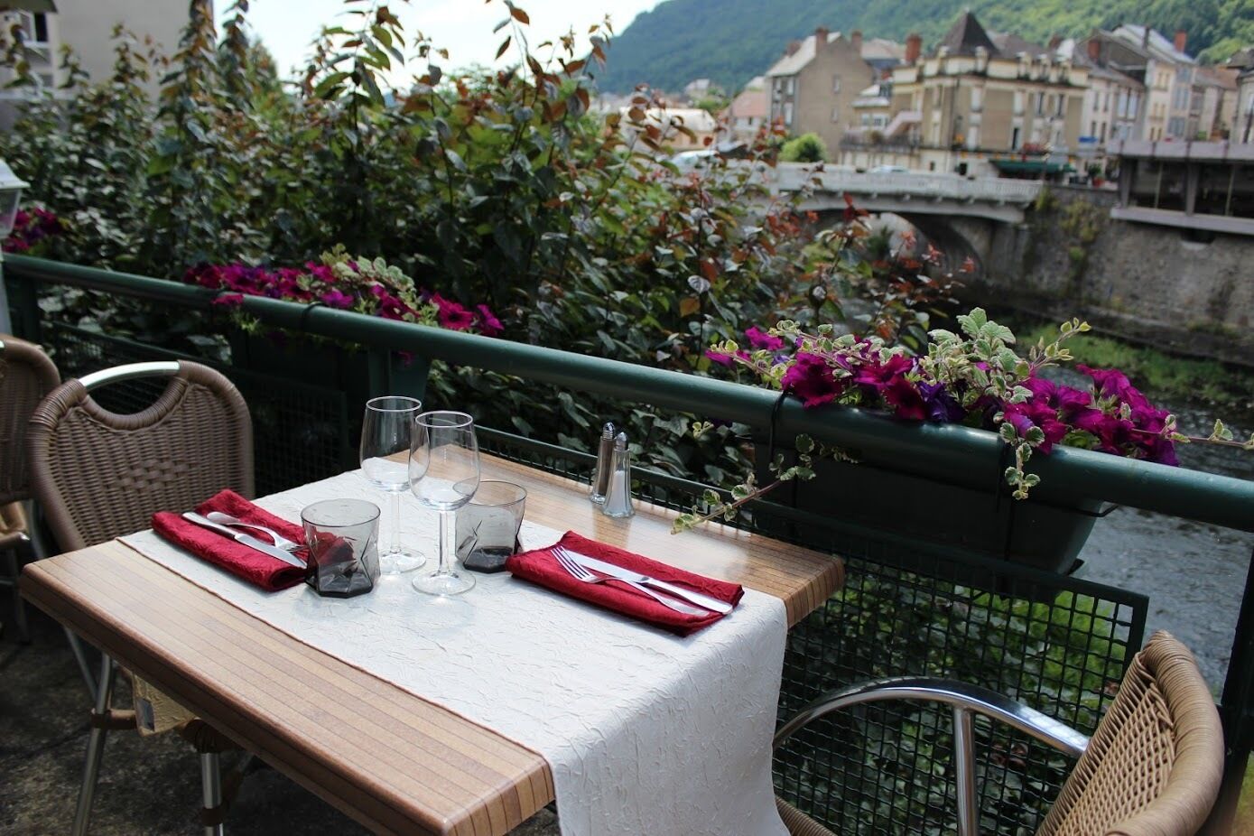 Un tavolo imbandito per due, con bicchieri da vino, un condimento e un tovagliolo rosso, su un balcone con vista su un fiume e degli edifici.