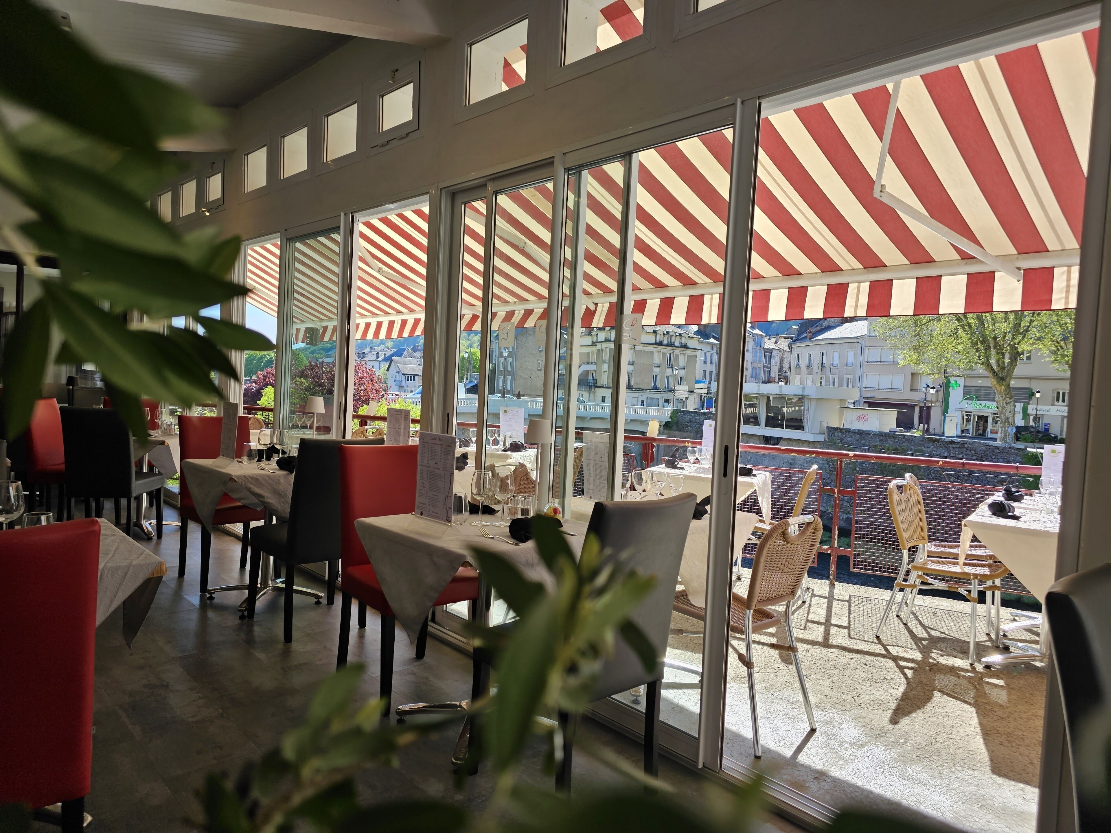 Un restaurante con amplios ventanales y toldos a rayas rojas, que ofrece vistas a edificios y un árbol.