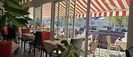 Un restaurante con amplios ventanales y toldos a rayas rojas, que ofrece vistas a edificios y un árbol.