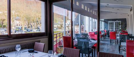 Un restaurante con mesas dispuestas para comer, grandes ventanales que ofrecen vistas a la calle y árboles, y una decoración interior moderna.