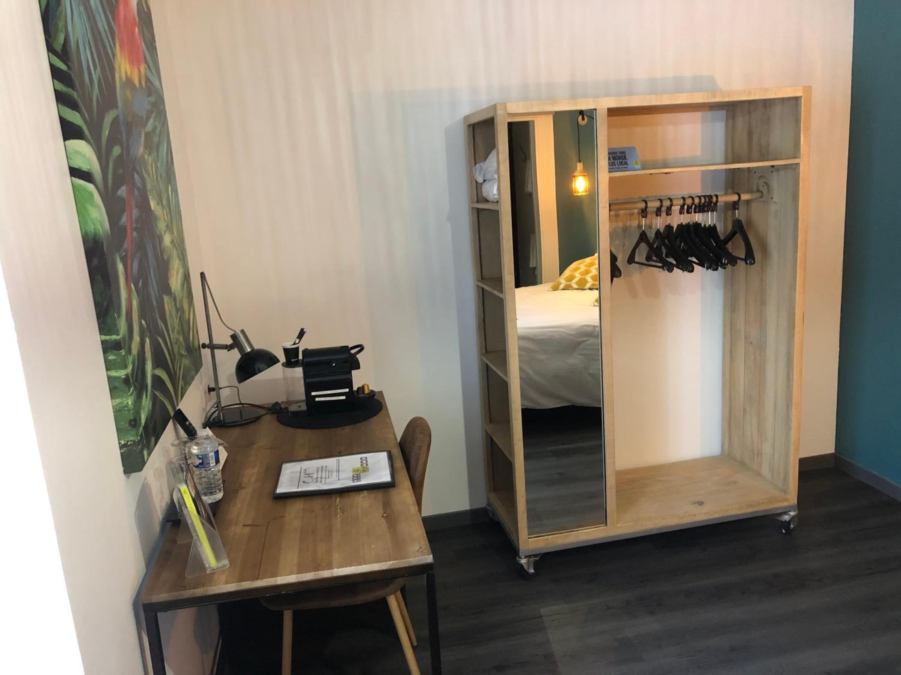 A small room with a wooden desk, a bed with a white and yellow bedspread, and a wooden wardrobe with a hanging rack.
