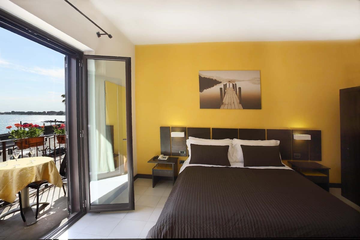 Une chambre d’hôtel comprenant un lit, une table de chevet, une commode, une fenêtre donnant sur la mer et un balcon avec une table et des chaises.