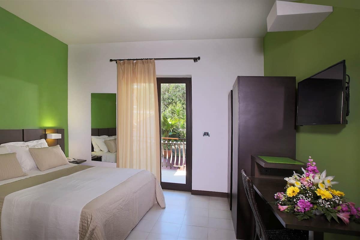 Une chambre d’hôtel avec un grand lit, un bureau avec une télévision, un balcon avec des rideaux et un mur végétal.