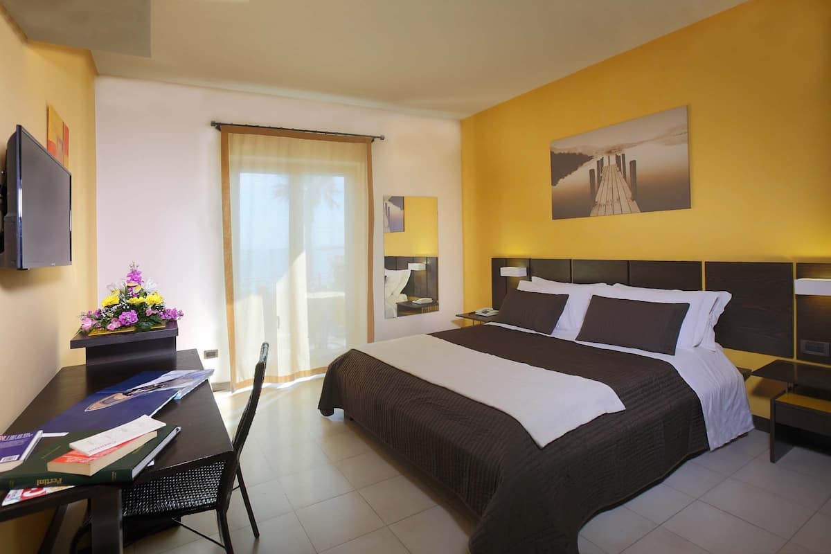 Une chambre d’hôtel avec un grand lit, un bureau avec des livres, une chaise, une télévision et une fenêtre avec des rideaux.