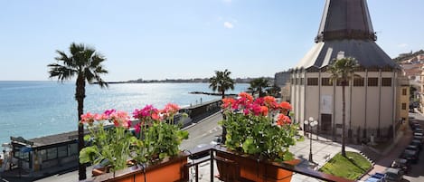 A balcony with potted flowers overlooking a coastal cityscape with a prominent church spire.