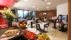 Een restaurant in een hotel met tafels, stoelen, een buffet met fruit en gebak, een televisie en een trap op de achtergrond.