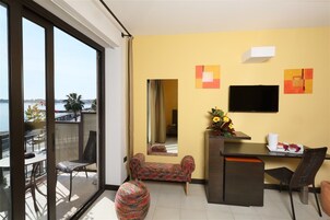 A hotel room with a balcony, a desk, a chair, a sofa, and a TV on the wall.