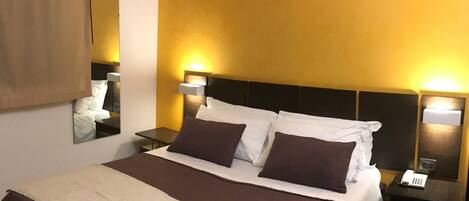 Habitación de hotel con una cama grande, dos mesitas de noche, un tocador, un espejo y una lámpara de pared.