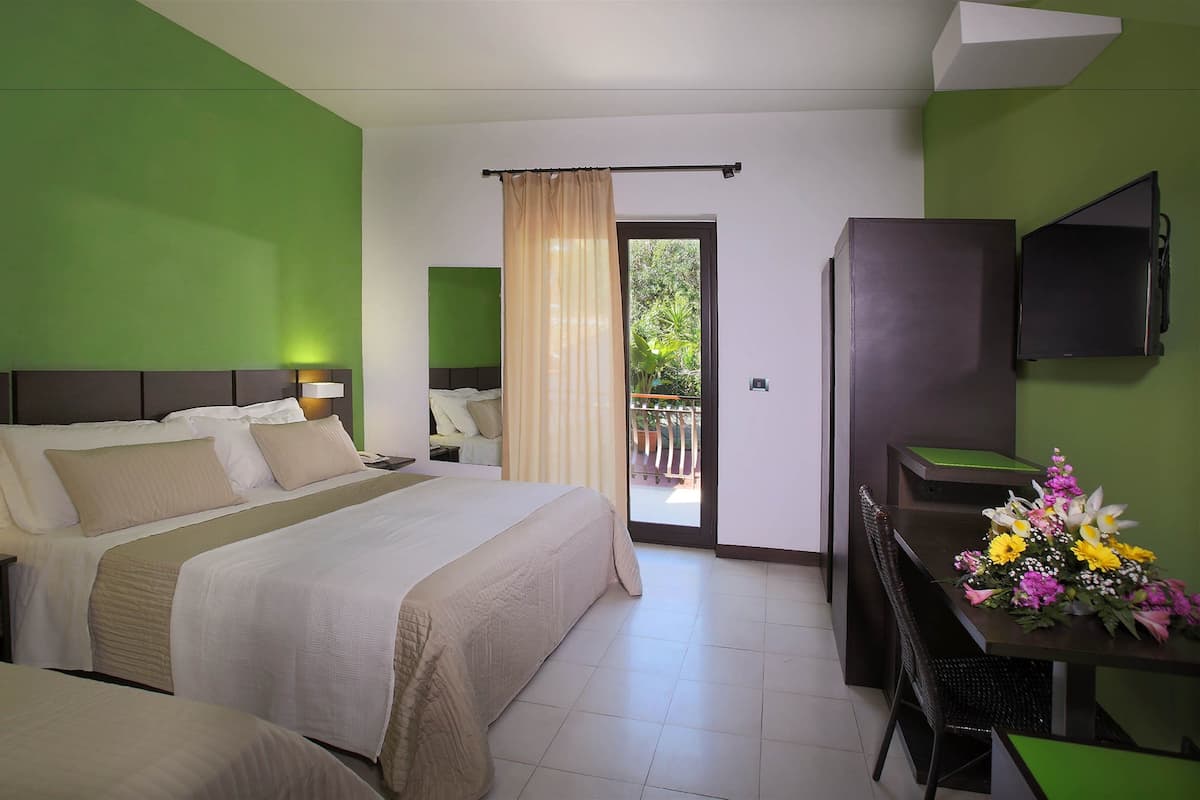 Une chambre d’hôtel équipée d’un lit, d’un bureau, d’une chaise, d’une télévision et d’un balcon donnant sur un paysage verdoyant.